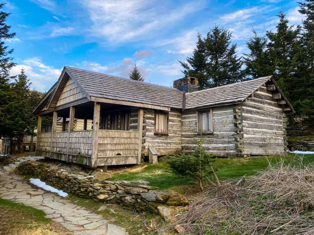 Rough-hewn log cabin with saddle-notched silver-gray logs, a shake roof, stone chimney, covered porch, and a flagstone path beside a low stone wall under a blue sky