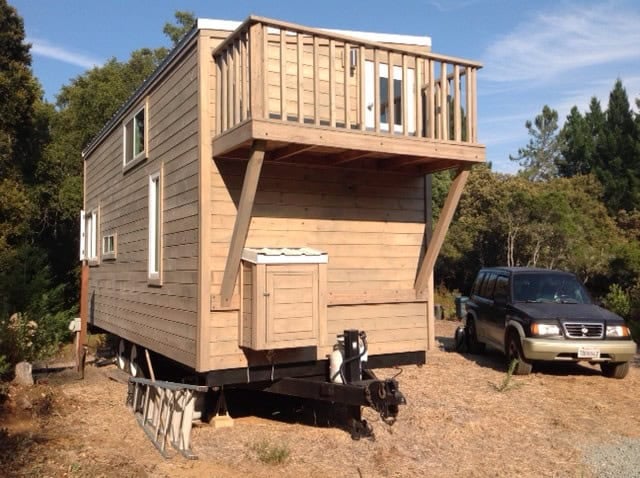 Side of the tan tiny house showing a small wood balcony on diagonal braces, white-trimmed windows, and the dual-axle trailer