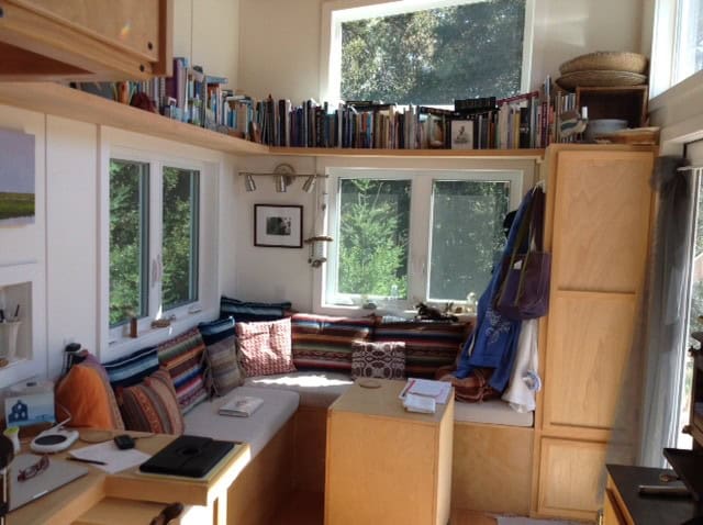 L-shaped built-in banquette with pillows under corner windows, a high wraparound bookshelf, and a tall wood cabinet in Lisa’s tiny house