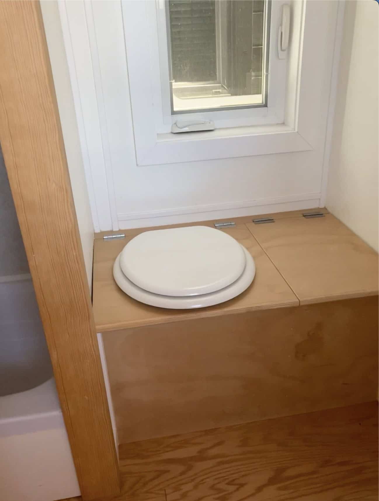 Composting toilet built into a light-wood bench with hinged lids under a small crank-out bathroom window