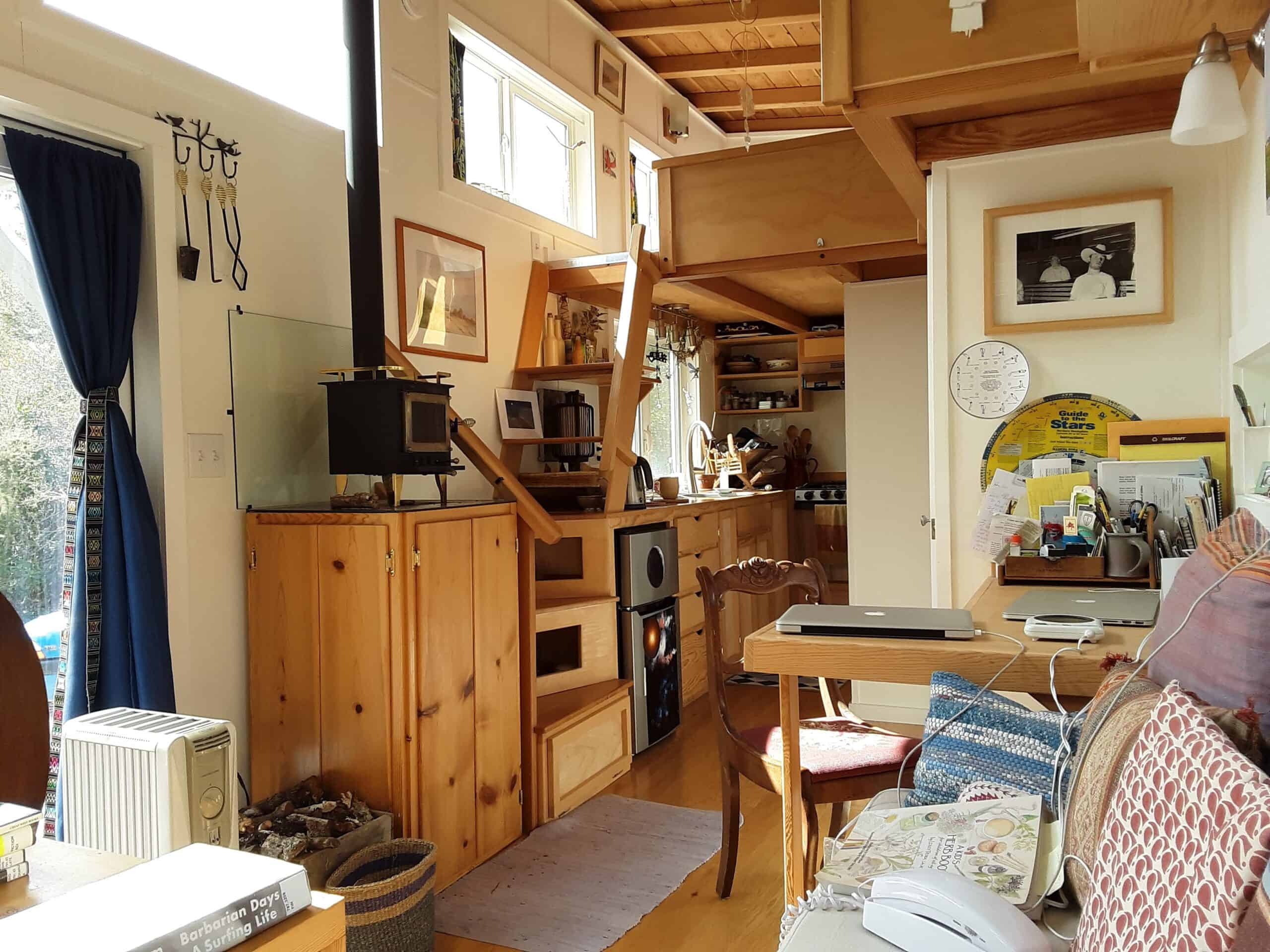 Looking down the light-wood interior of Lisa’s tiny house: banquette and desk in the foreground, a small wood stove, storage stairs, galley kitchen, and a loft above