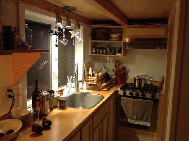 L-shaped tiny-house kitchen at night with a stainless sink under a window, wood counters, open shelves, and a compact four-burner gas range and oven