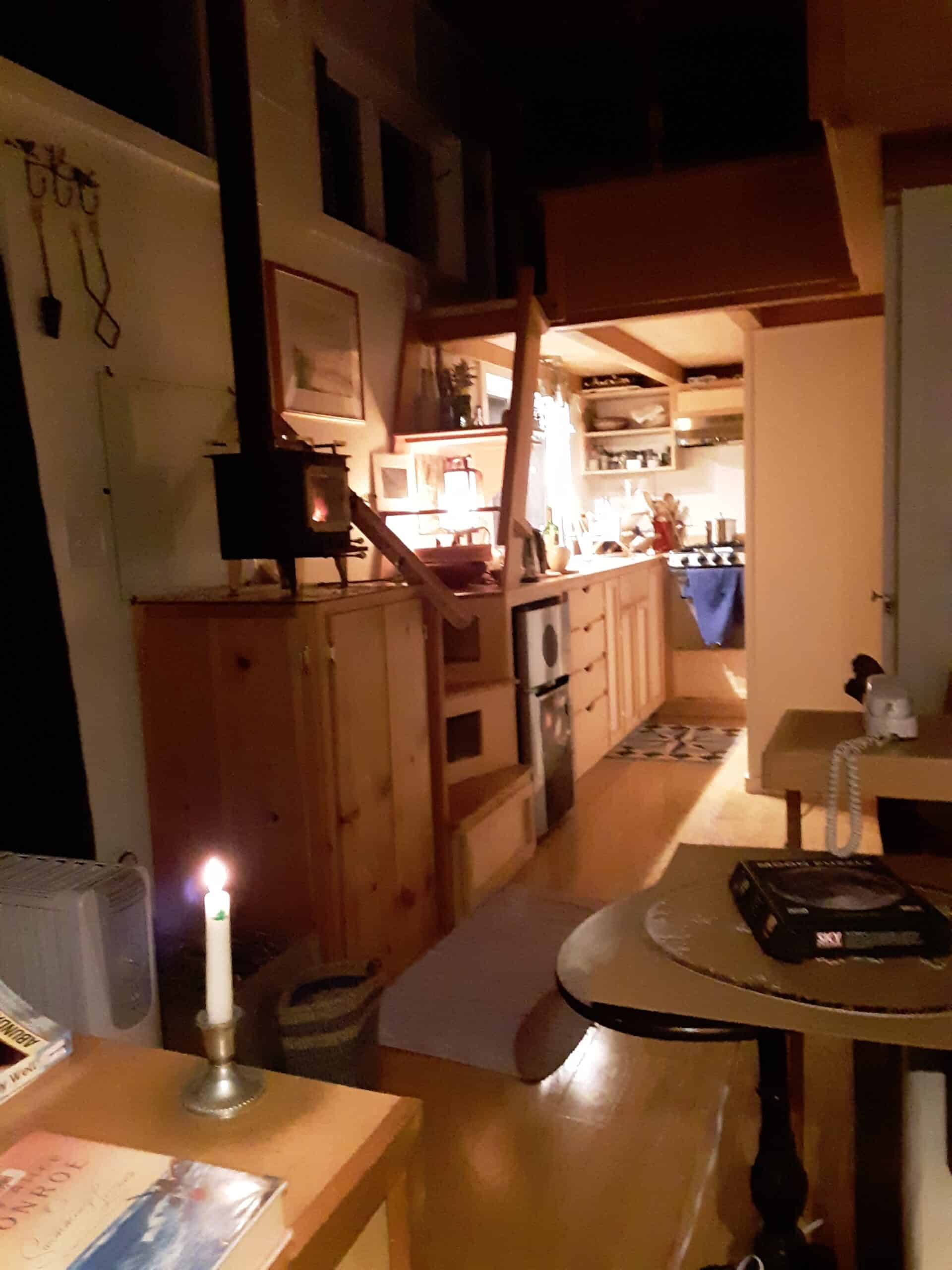 Warm nighttime view down the length of the tiny house: wood stove with a fire going, storage stairs, and a softly lit galley kitchen under the loft