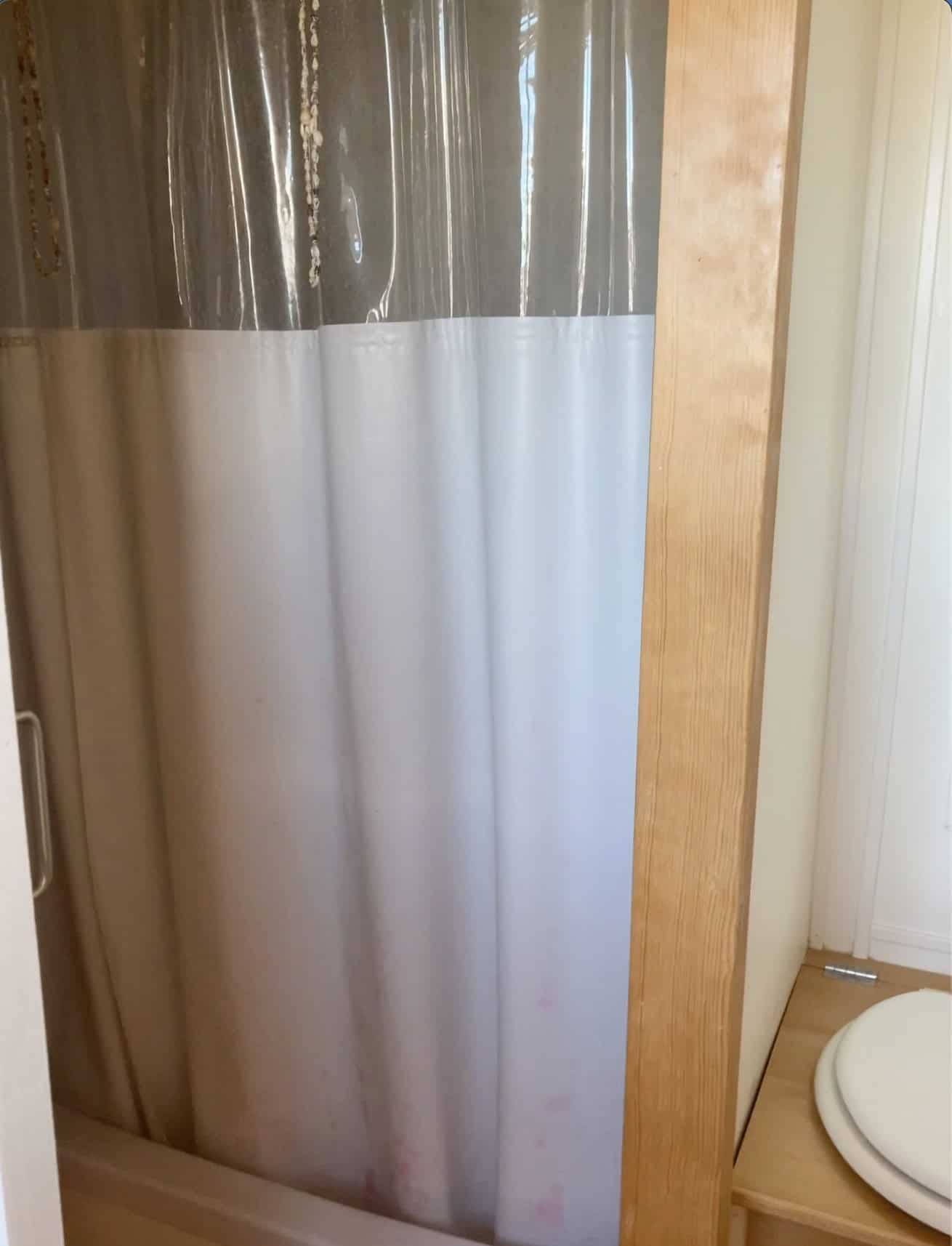 Compact shower stall framed in light wood with a clear-and-white curtain, next to the composting-toilet bench
