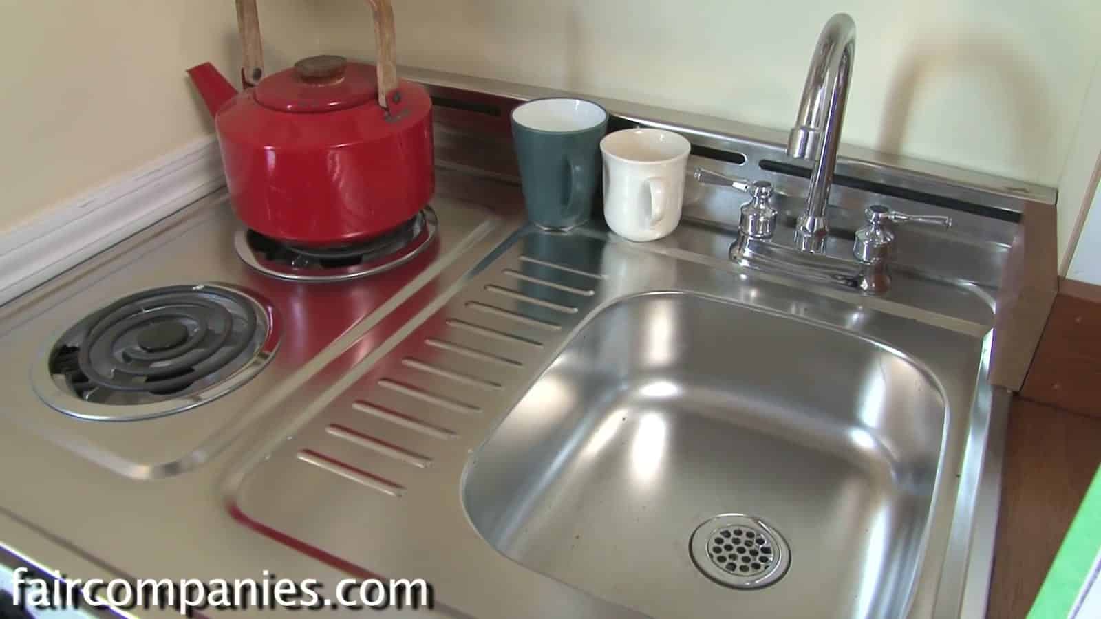 Compact stainless kitchenette with a two-burner electric cooktop and sink in the Little House on a Trailer