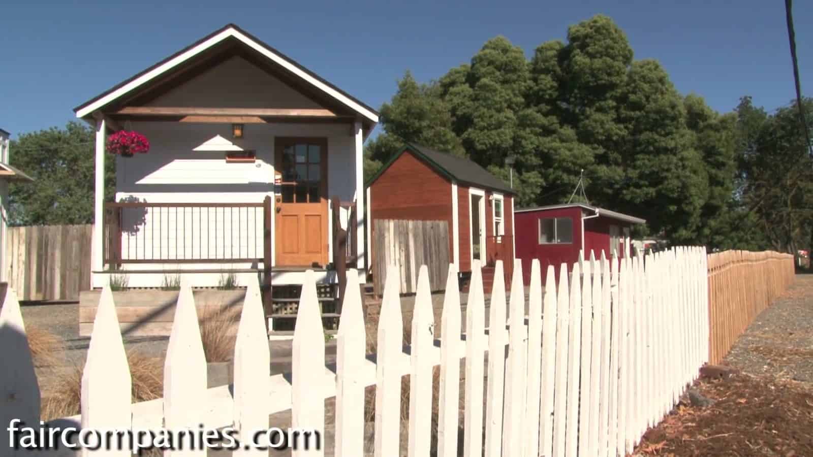 Little House on a Trailer behind a white picket fence beside other small houses on a sales lot