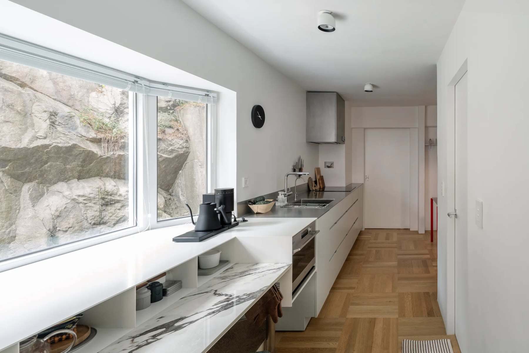 Long white breakfast bar under a wide window facing a natural rock wall, with the kitchen beyond