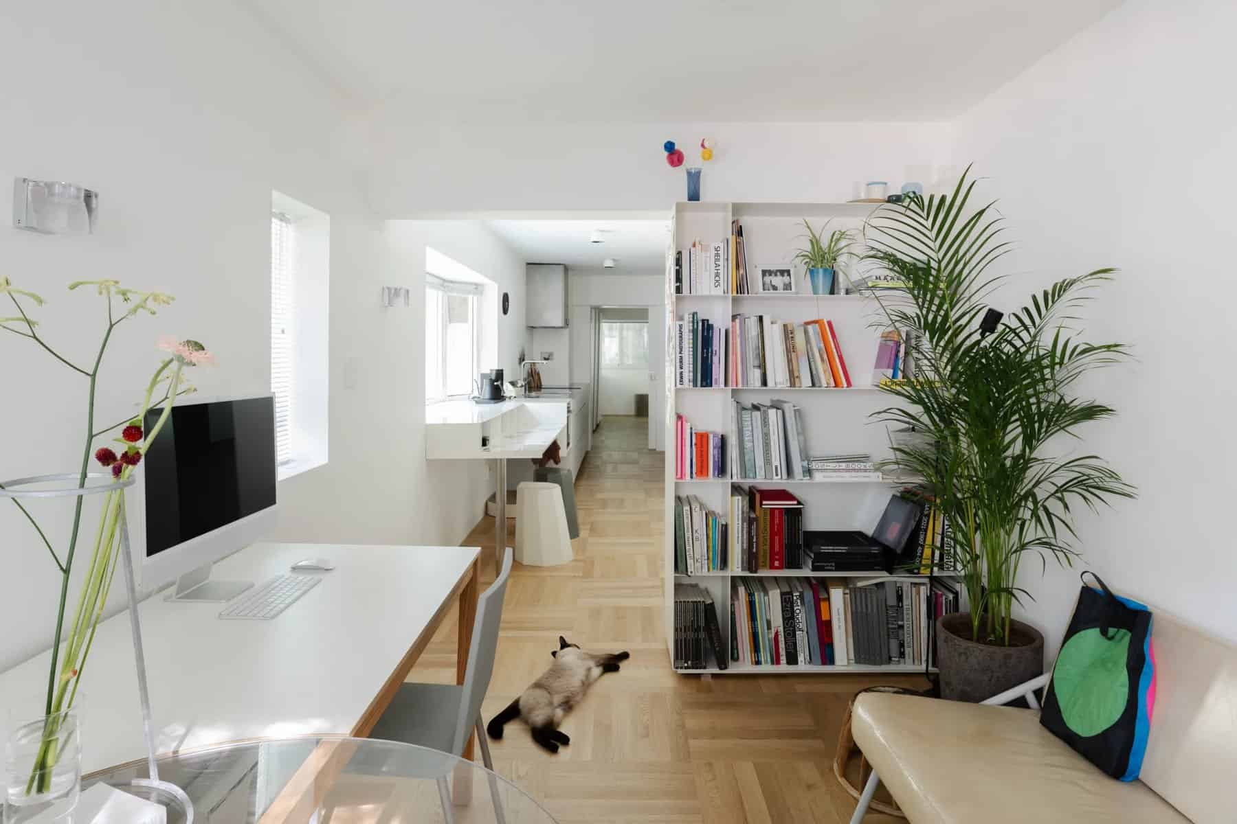 Bright living room with a white desk and computer, a tall bookshelf, a plant, and a view down the length of the apartment