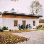 White corrugated shipping-container tiny house with a dark shed roof, wood-shingle band, black-framed glass door, two rocking chairs, and gravel in front — Luna Cottage in Hocking Hills