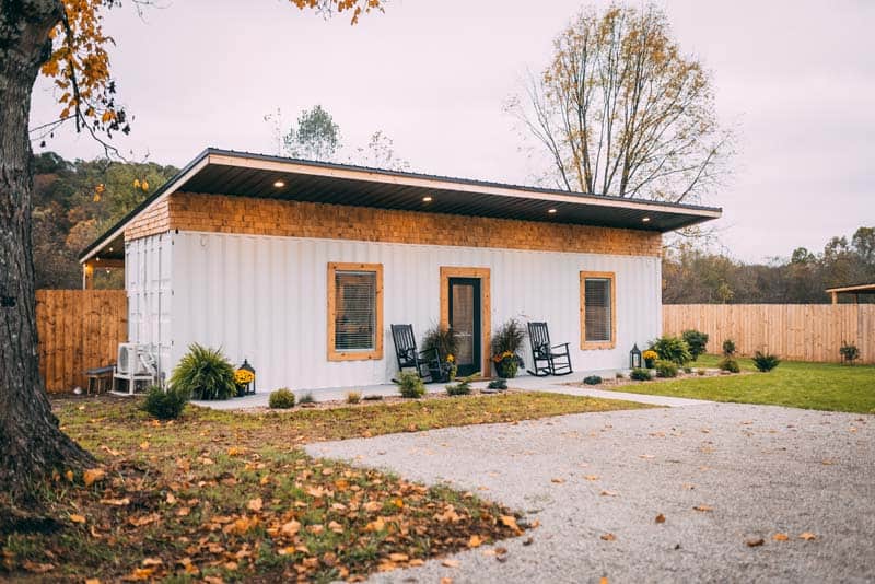 White corrugated shipping-container tiny house with a dark shed roof, wood-shingle band, black-framed glass door, two rocking chairs, and gravel in front — Luna Cottage in Hocking Hills