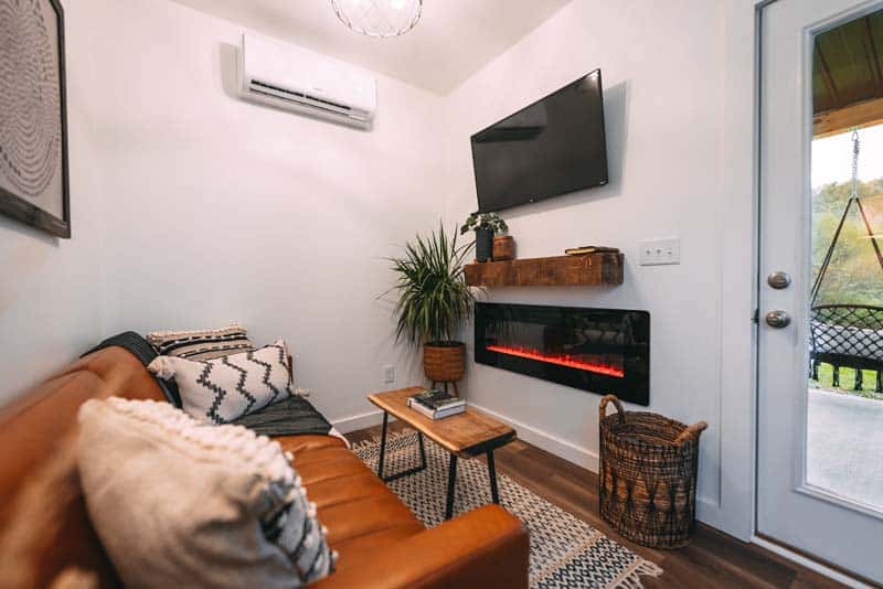 Living room of Luna Cottage: tan leather sofa, live-edge coffee table, wall-mounted TV over a fireplace, glass door open to a macrame porch swing
