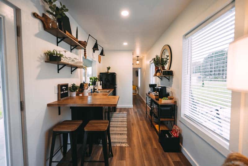 Kitchen aisle of Luna Cottage: butcher-block breakfast bar with two stools, wood shelves over a sink, black refrigerator, and a coffee-bar cart along a white container-width hall