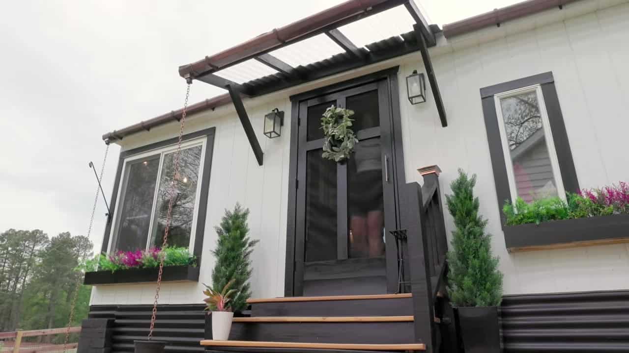 White board-and-batten 12x20 shed tiny house with black trim, screen door wreath, flower boxes, and wooden steps in Central Virginia