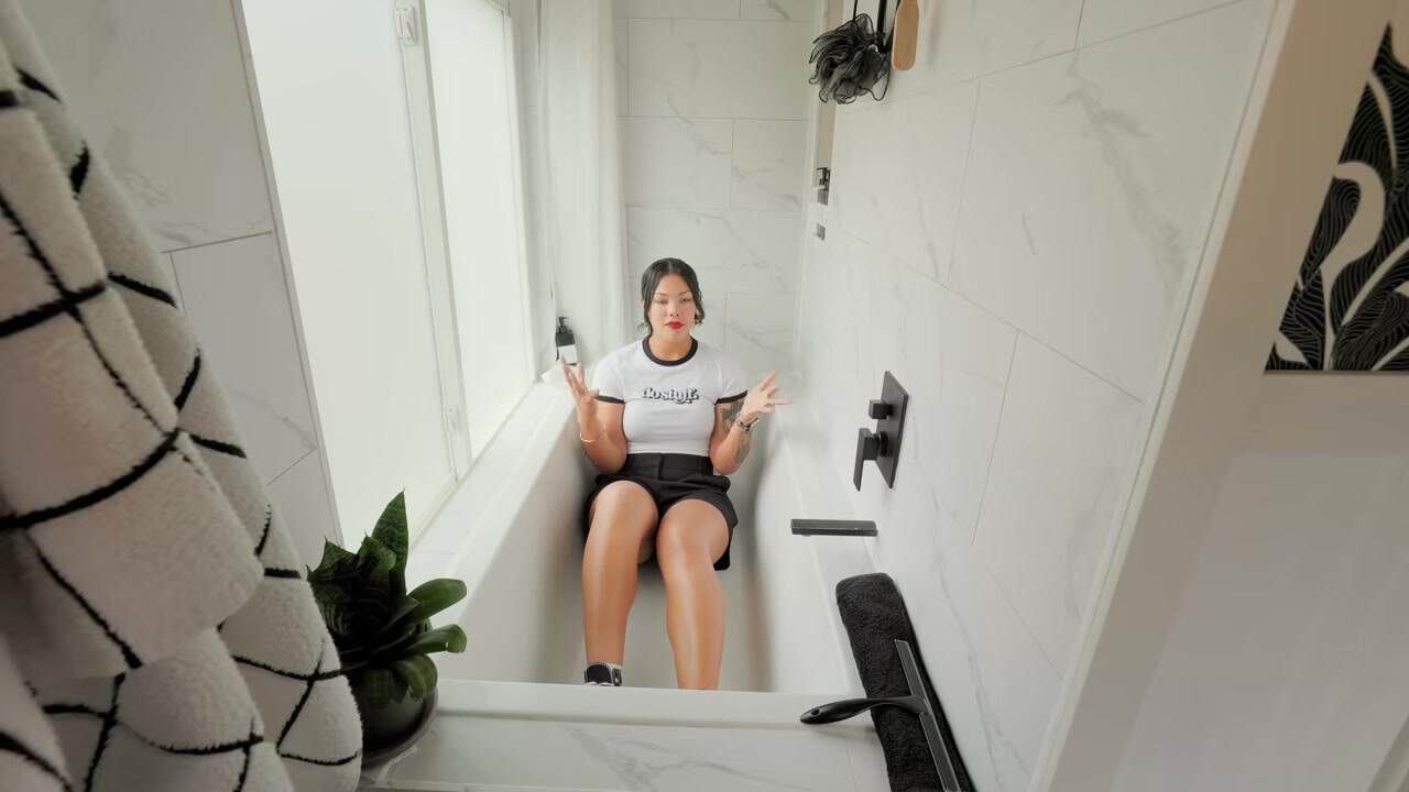 Margo sitting in a full-size white alcove bathtub with marble-look tile and matte-black fixtures to show the scale