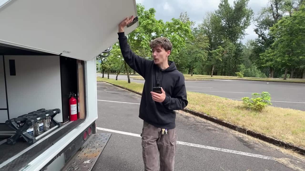 Maxwell Peters holding open an exterior storage hatch on his moving van tiny home with tanks and fire extinguisher inside