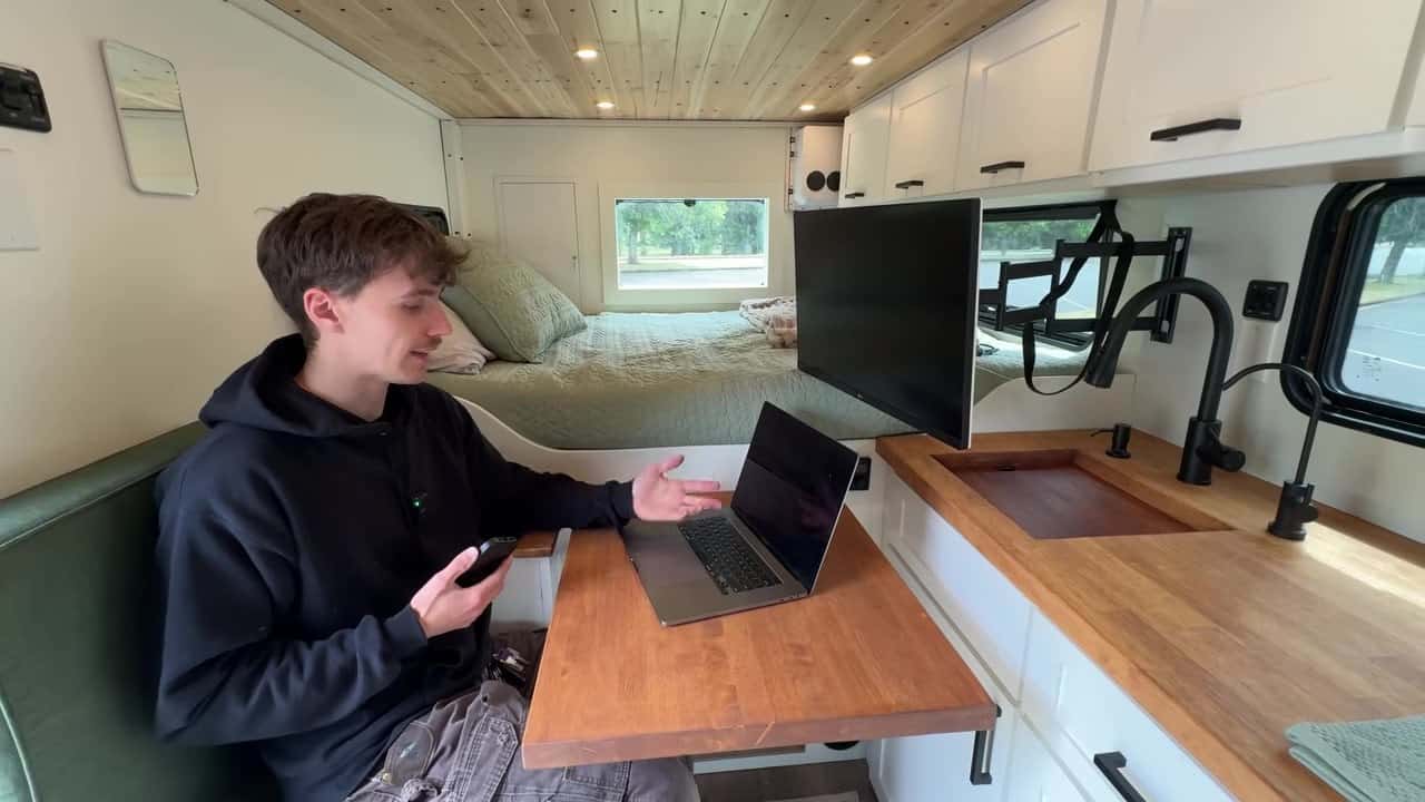 Maxwell Peters at the wood dinette workspace with swivel monitor and sage-green bed behind him in his tiny home van