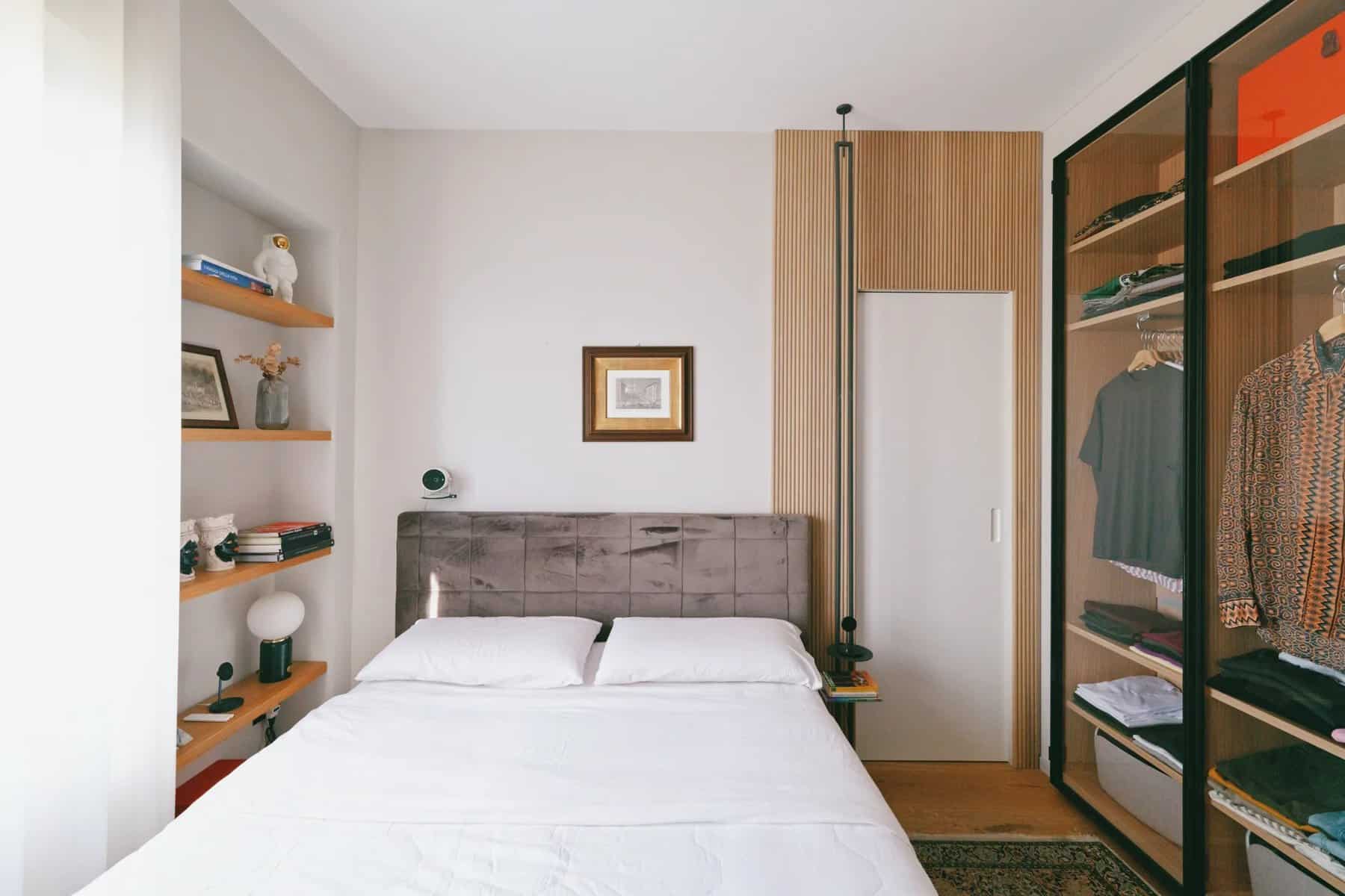 Bedroom with a grey upholstered bed, wood wall shelves, and a glass-front wardrobe