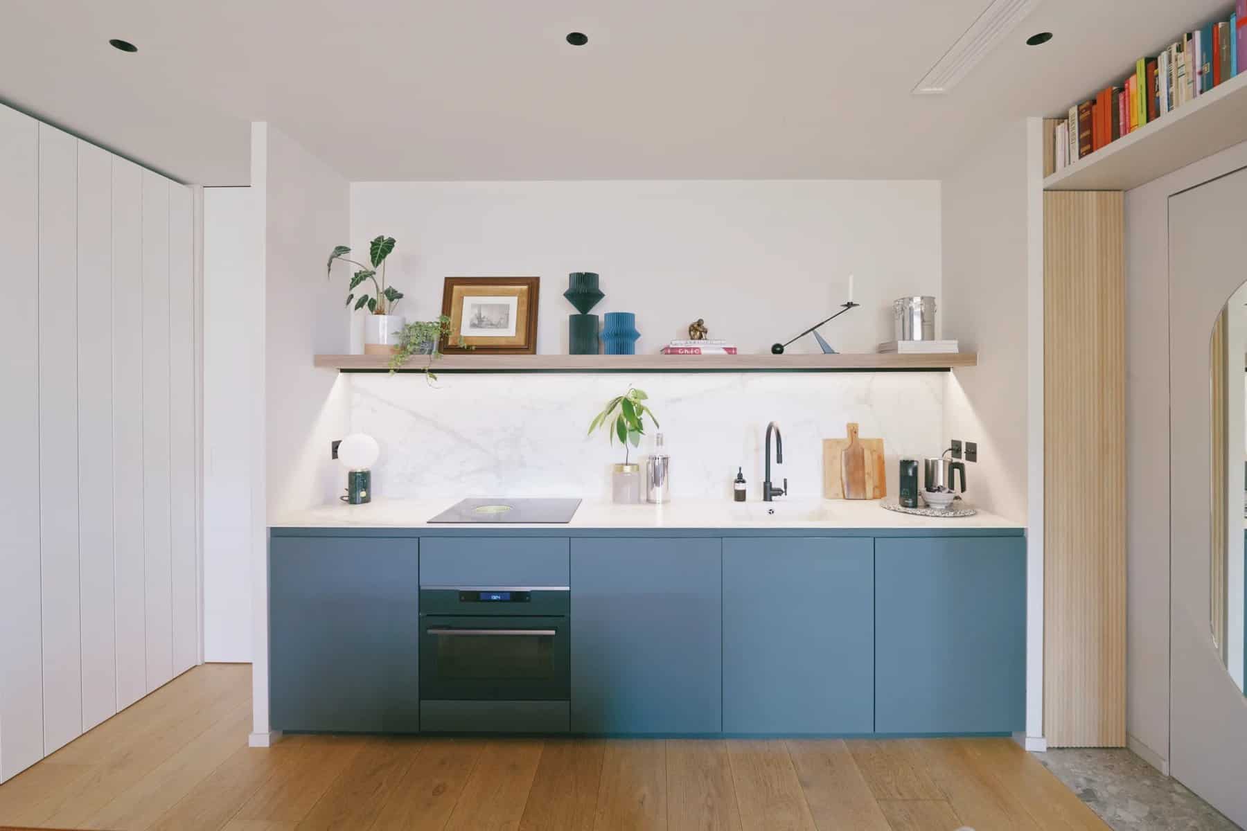 Minimal kitchen with dark blue cabinets, white stone counter and backsplash, and a single wood shelf