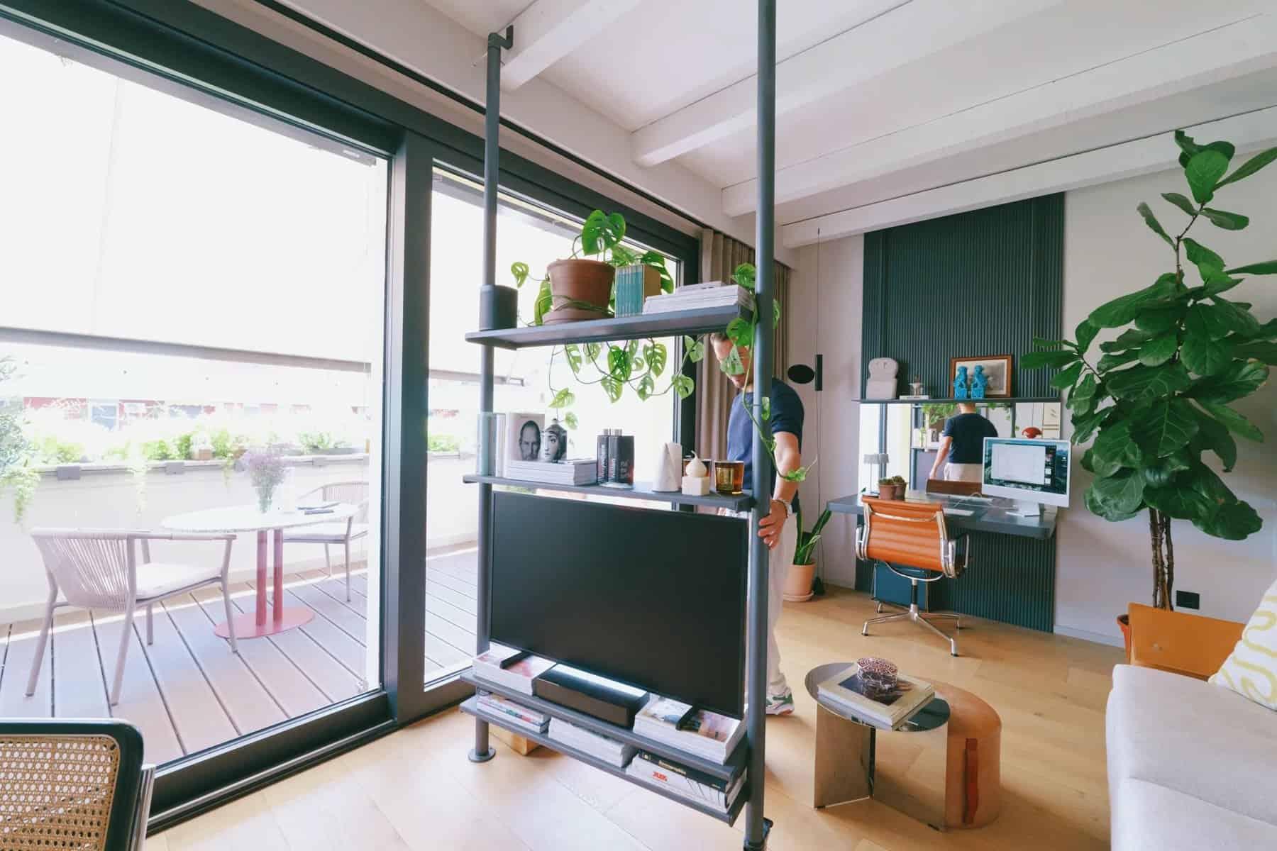 Rotating metal shelving unit holding a TV and plants, dividing the living room from the desk area