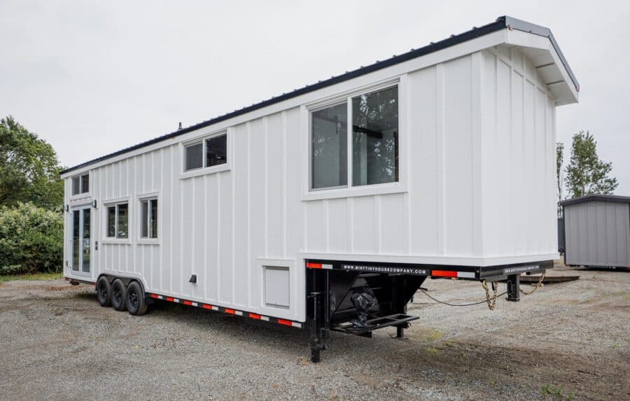 Mint Arctic tiny house on a triple-axle trailer: white board-and-batten, grey metal roof, French doors, gooseneck over the hitch, minttinyhousecompany.com on the frame, gravel lot