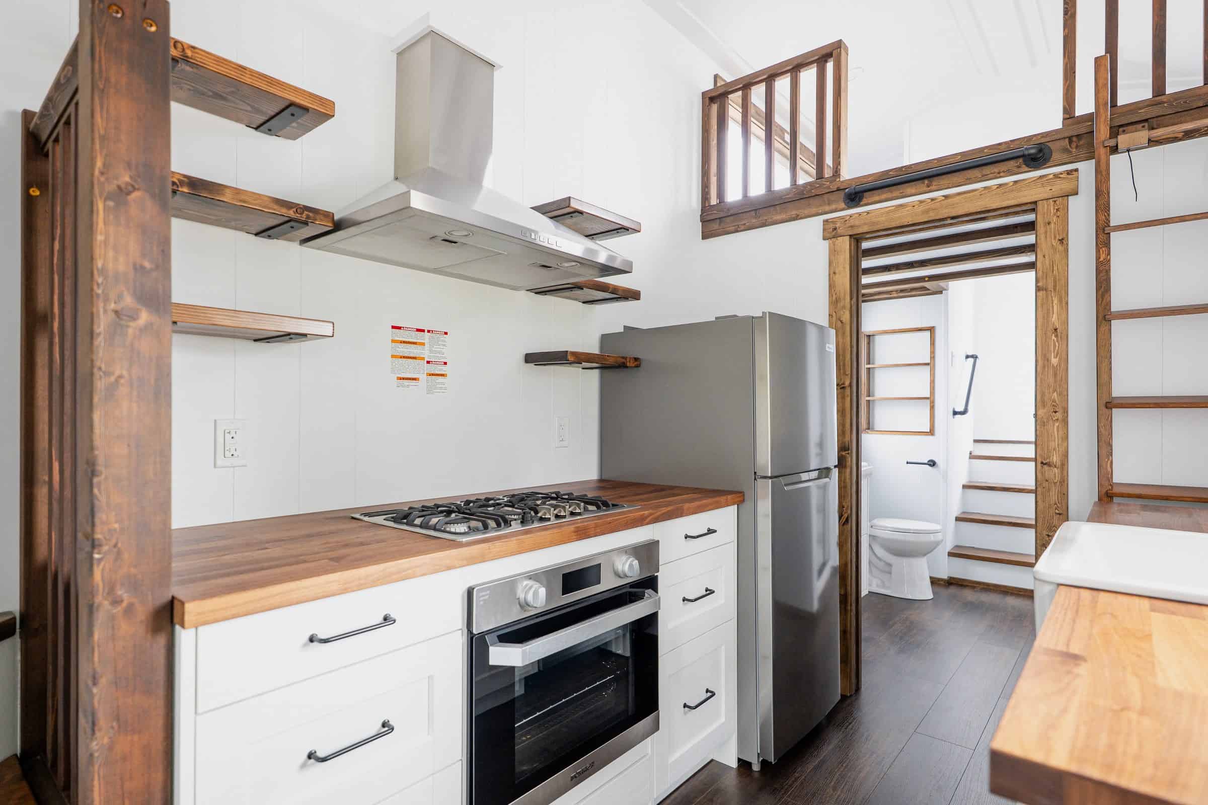 Arctic kitchen: butcher-block, propane cooktop and oven, stainless range hood and 24-inch fridge, doorway to stairs and bath, loft rail above