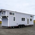 Mint Loft Ruby tiny house on a triple-axle black trailer: light grey vertical siding, white trim and glass door, dark roof, cantilevered loft over the tongue, parked on gravel