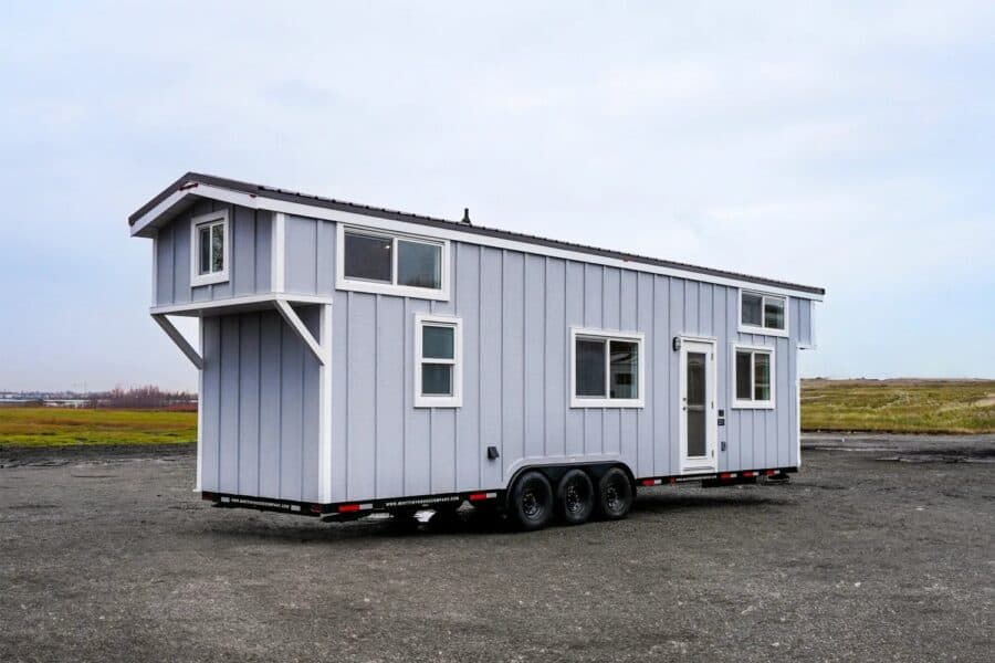 Mint Loft Ruby tiny house on a triple-axle black trailer: light grey vertical siding, white trim and glass door, dark roof, cantilevered loft over the tongue, parked on gravel