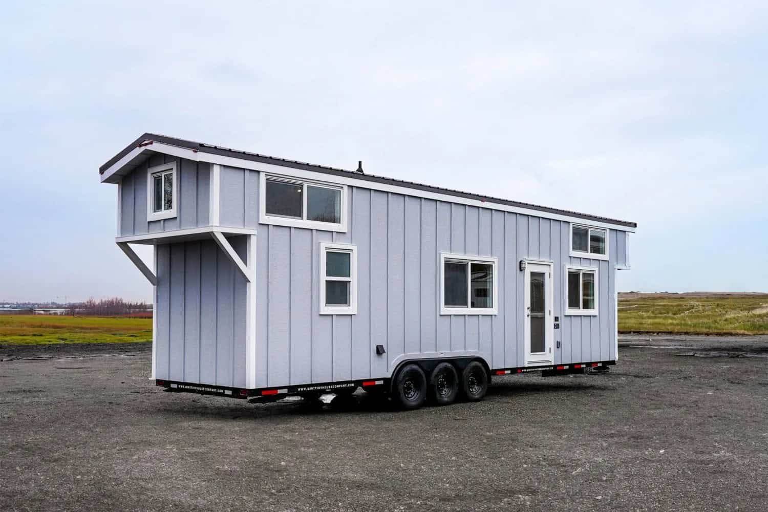 Mint Loft Ruby tiny house on a triple-axle black trailer: light grey vertical siding, white trim and glass door, dark roof, cantilevered loft over the tongue, parked on gravel