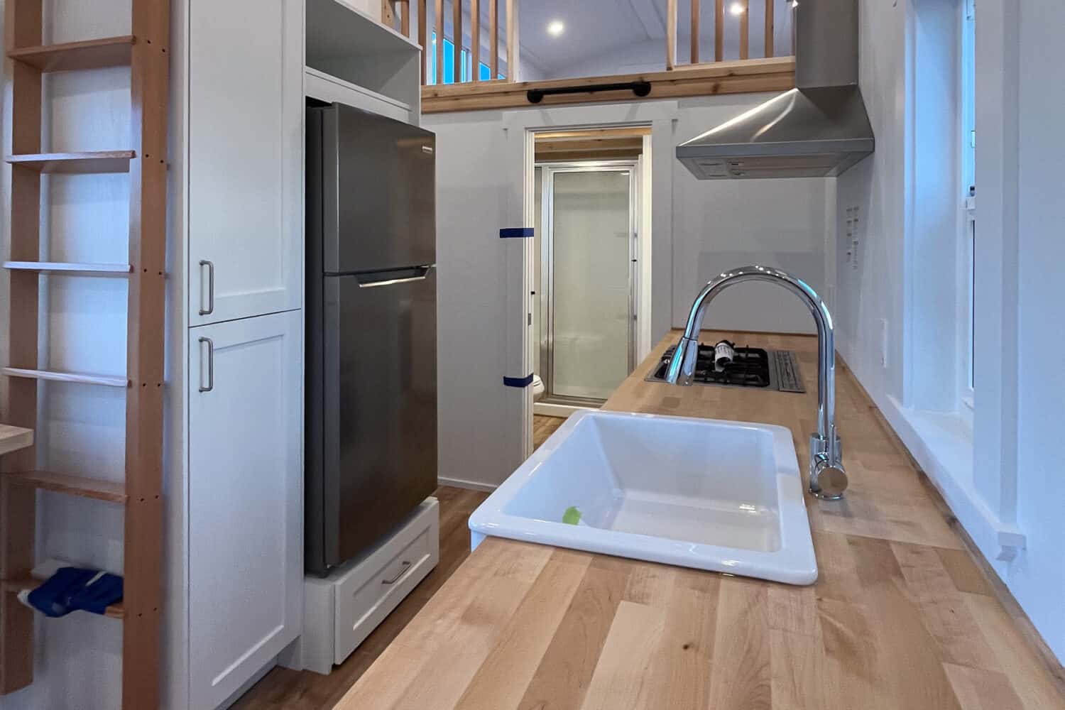 Kitchen looking toward the loft and bath: butcher-block, apron sink, cooktop and range hood, stainless fridge, wood stairs, glass shower door at the end of the hall