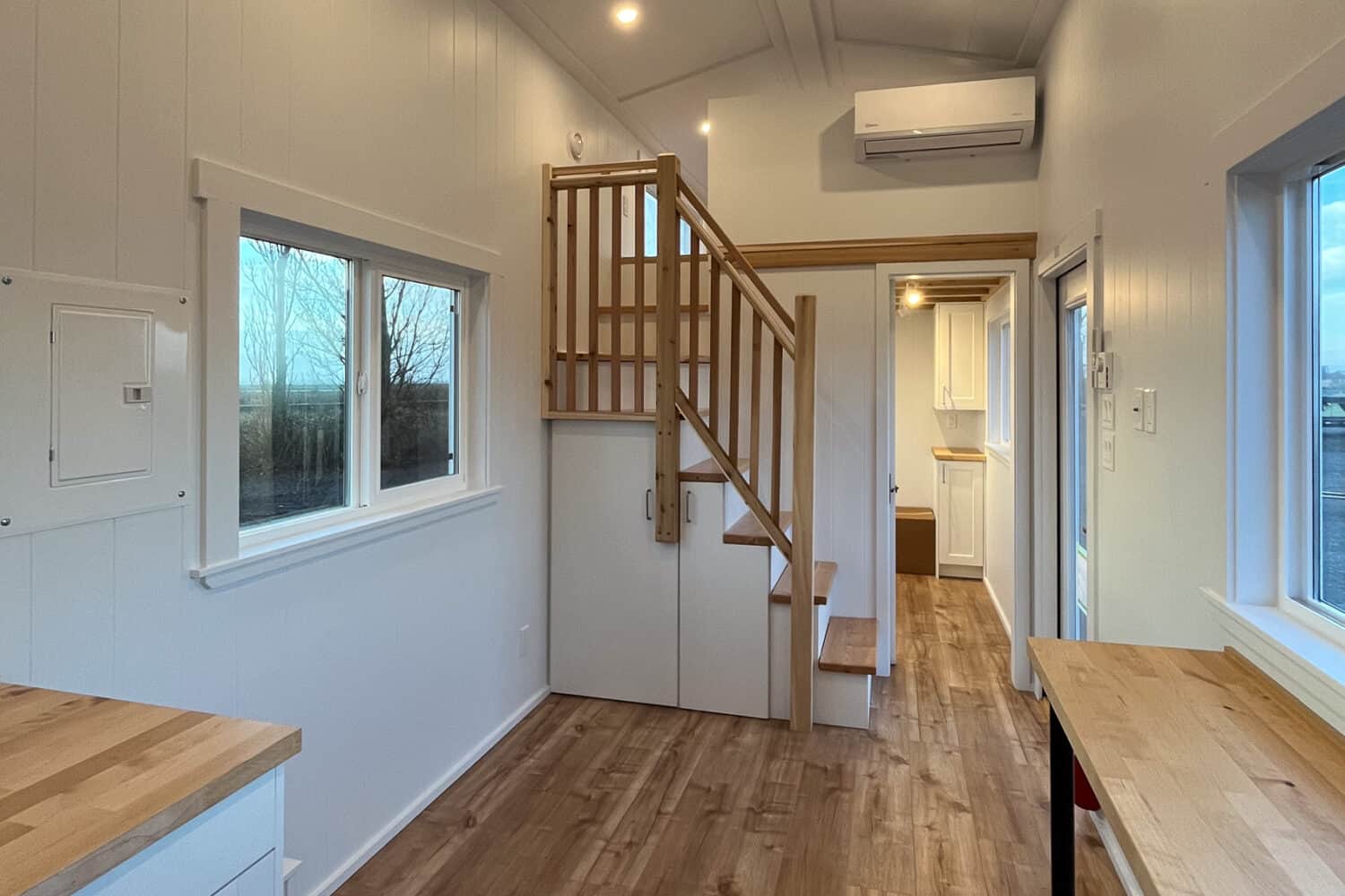 Living run of the Loft Ruby: wood storage stairs with white cabinets in the stringer, vaulted white ceiling, mini-split over a rear doorway, light plank floor