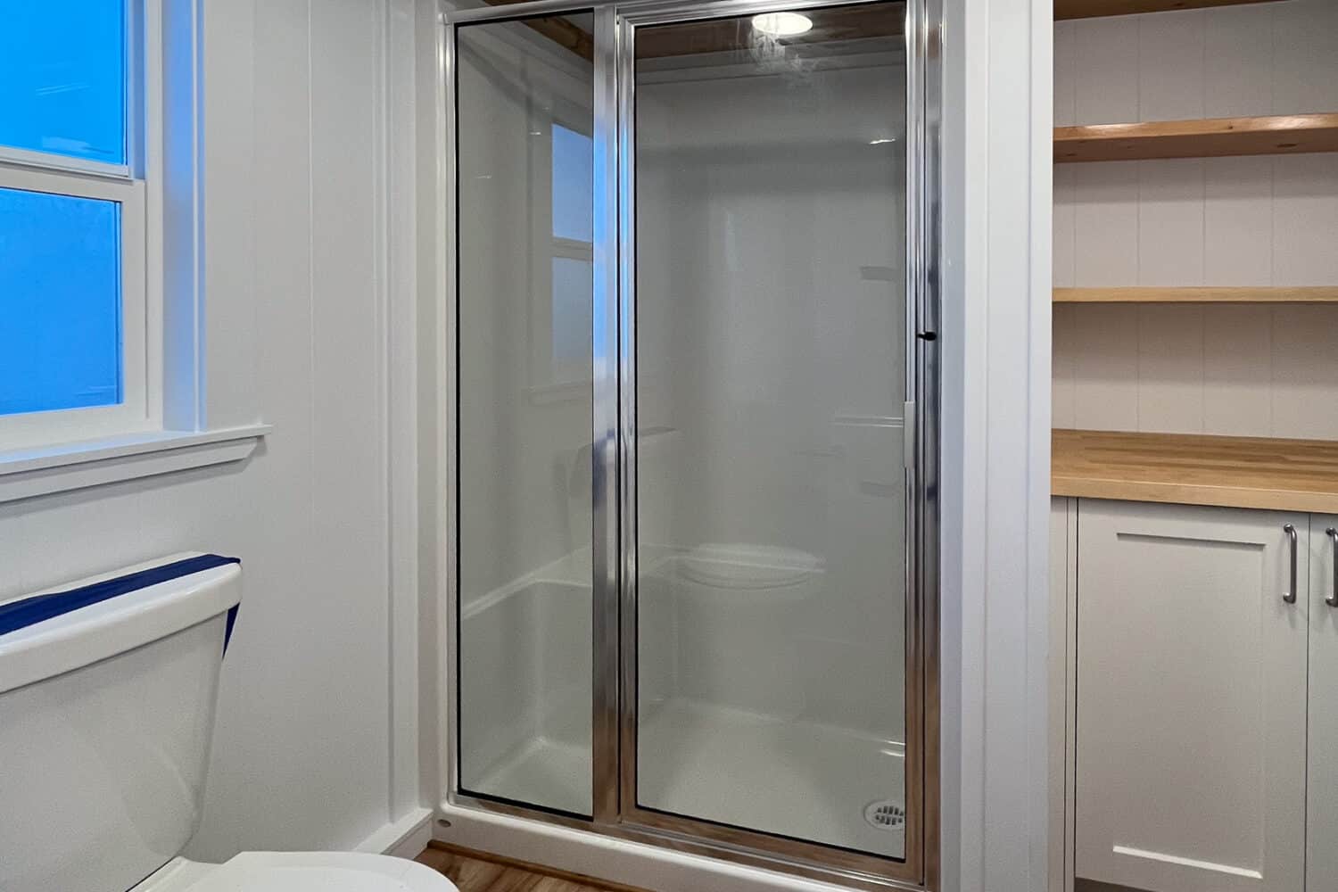 48x32 shower with chrome-framed glass door and a molded seat, white flush toilet, wood shelves beside the stall