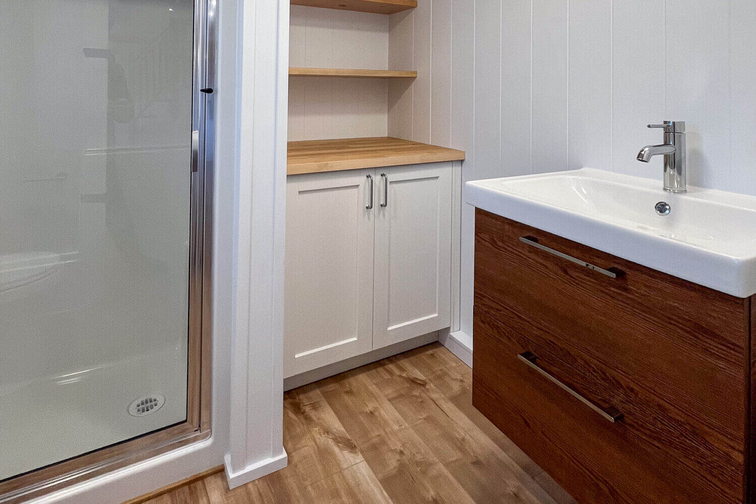 Bathroom vanity: dark-wood floating drawers, white rectangular sink, chrome faucet, white cabinet and two wood shelves, shower glass at left