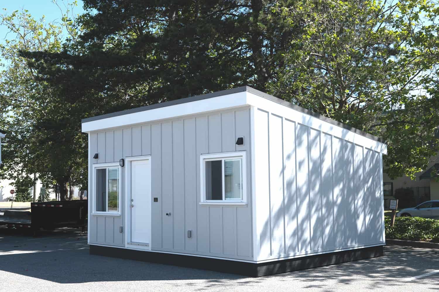 Three-quarter view of the Whirlwind Grey modular ADU: white entry door, white fascia and corner boards, dark skid base on asphalt, trees behind