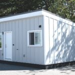 Whirlwind Grey modular ADU on an asphalt lot in Delta BC: white door and trim, white-framed sliders, dark base, trees behind — not a tiny house on wheels