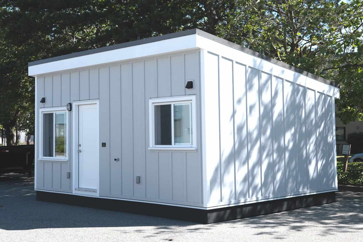 Whirlwind Grey modular ADU on an asphalt lot in Delta BC: white door and trim, white-framed sliders, dark base, trees behind — not a tiny house on wheels