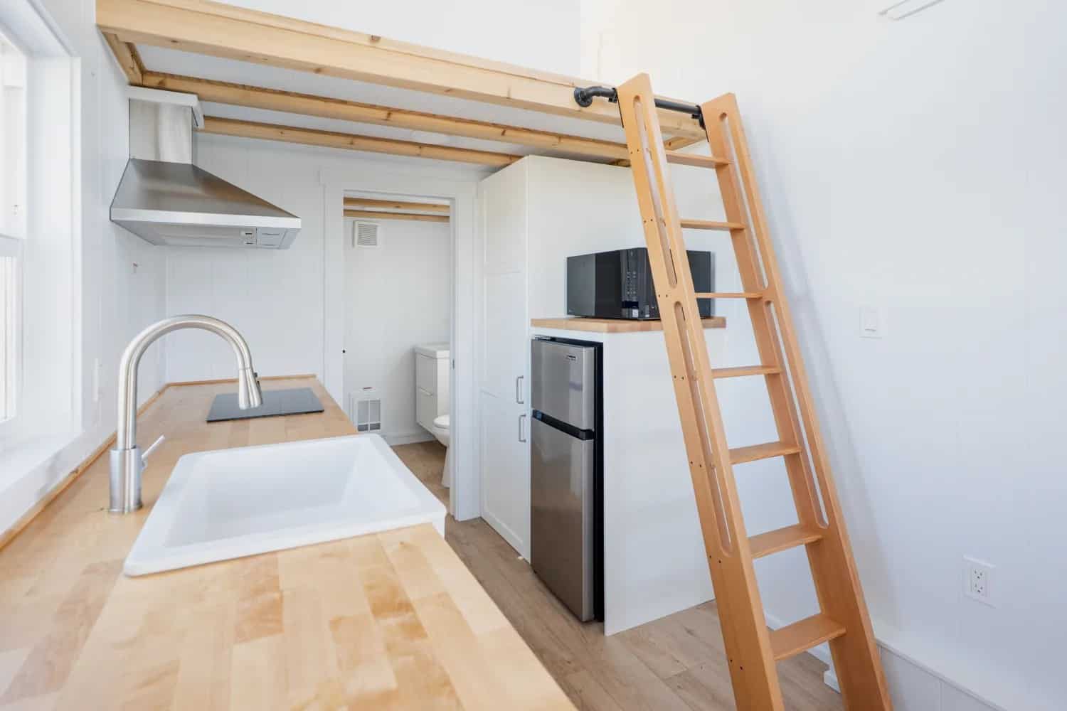 From the galley toward the bath: butcher-block counter, stainless mini fridge, wood loft ladder, open doorway with a white toilet and vanity