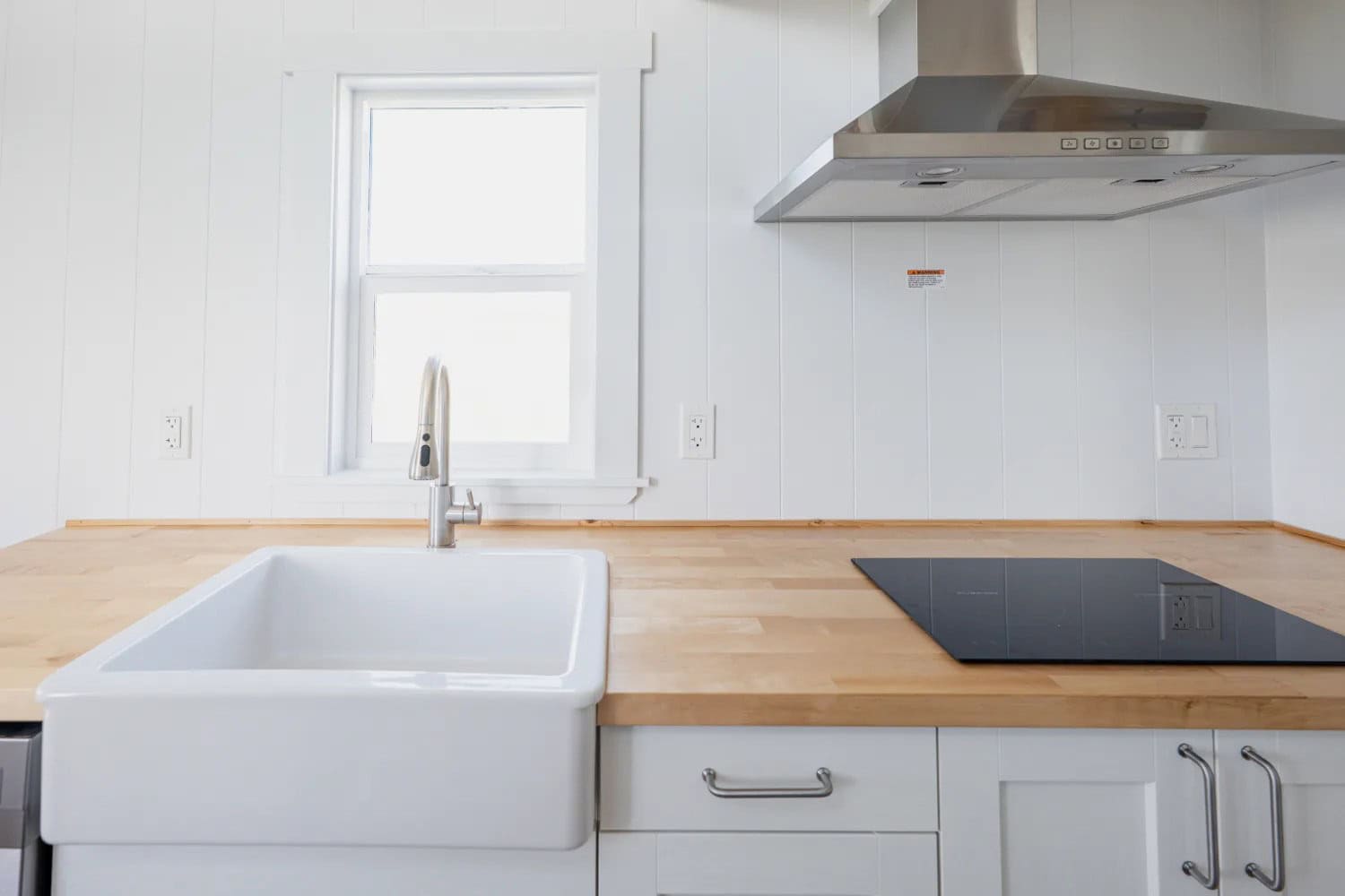 Close kitchen: light butcher-block veneer, white ceramic apron sink, pull-out faucet, black two-burner electric cooktop, stainless range hood, white shaker drawers