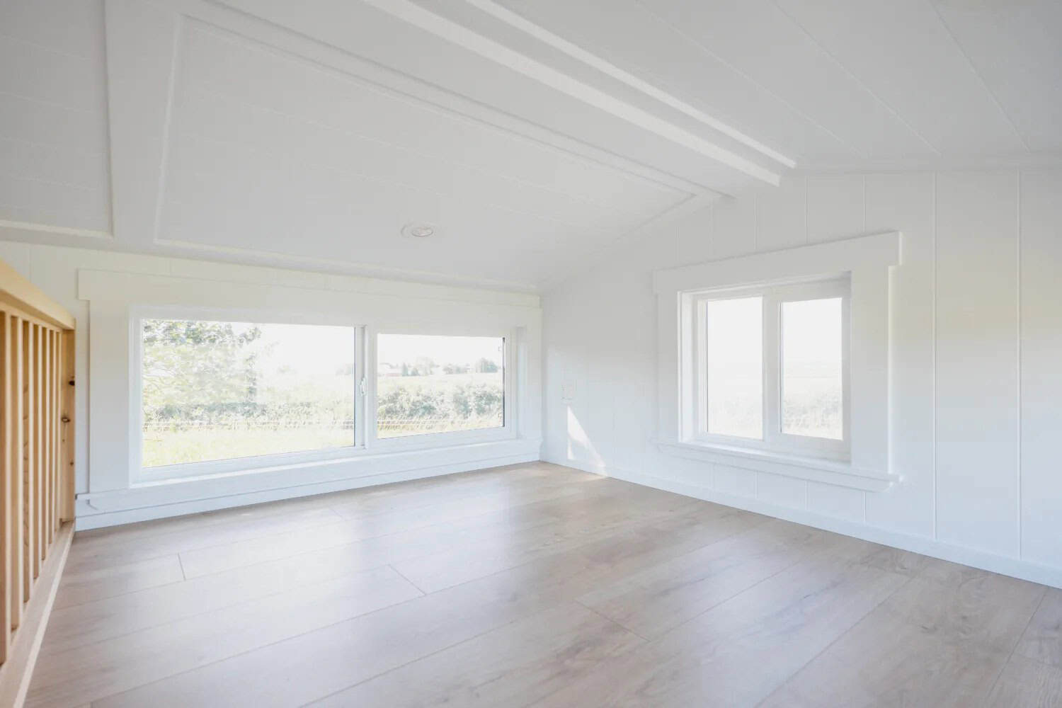 Empty sleeping loft: white wall siding, light plank floor, wide slider, smaller side window, wood railing at the loft edge