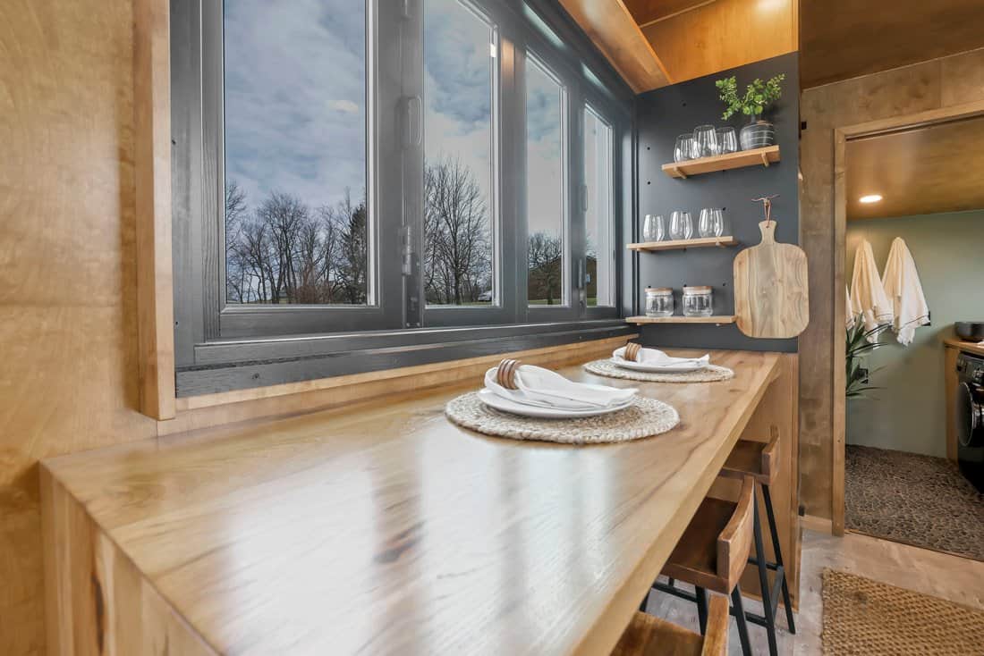 Inside the Modern Tiny Living Catalina: long wood bar counter under a wall of black-framed windows, with open shelves and stools