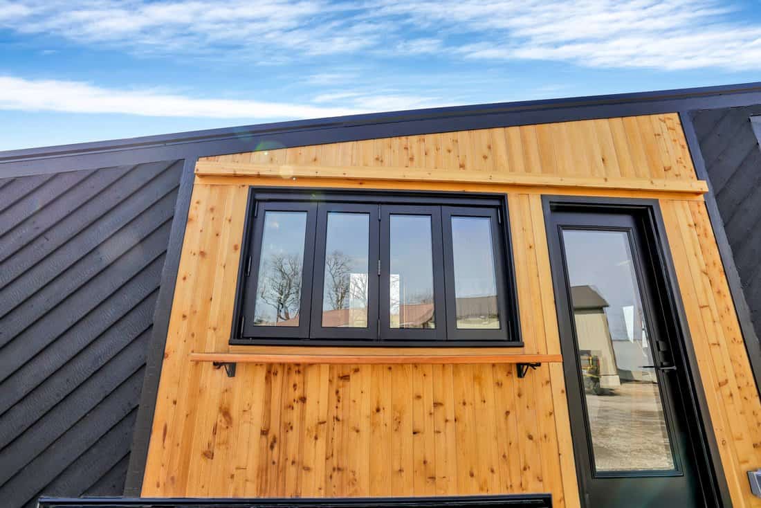 Cedar-clad bump-out on the Modern Tiny Living Catalina with a row of black-framed bar windows over an exterior ledge, next to the glass entry door