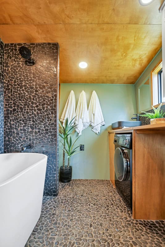 Upgraded Catalina bathroom: freestanding soaking tub, black pebble-tile shower wall and floor, washer-dryer under a wood vanity