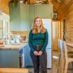 Single-story tiny cottage kitchen and living room, vaulted wood ceiling, sage cabinets, Mollie standing in the center