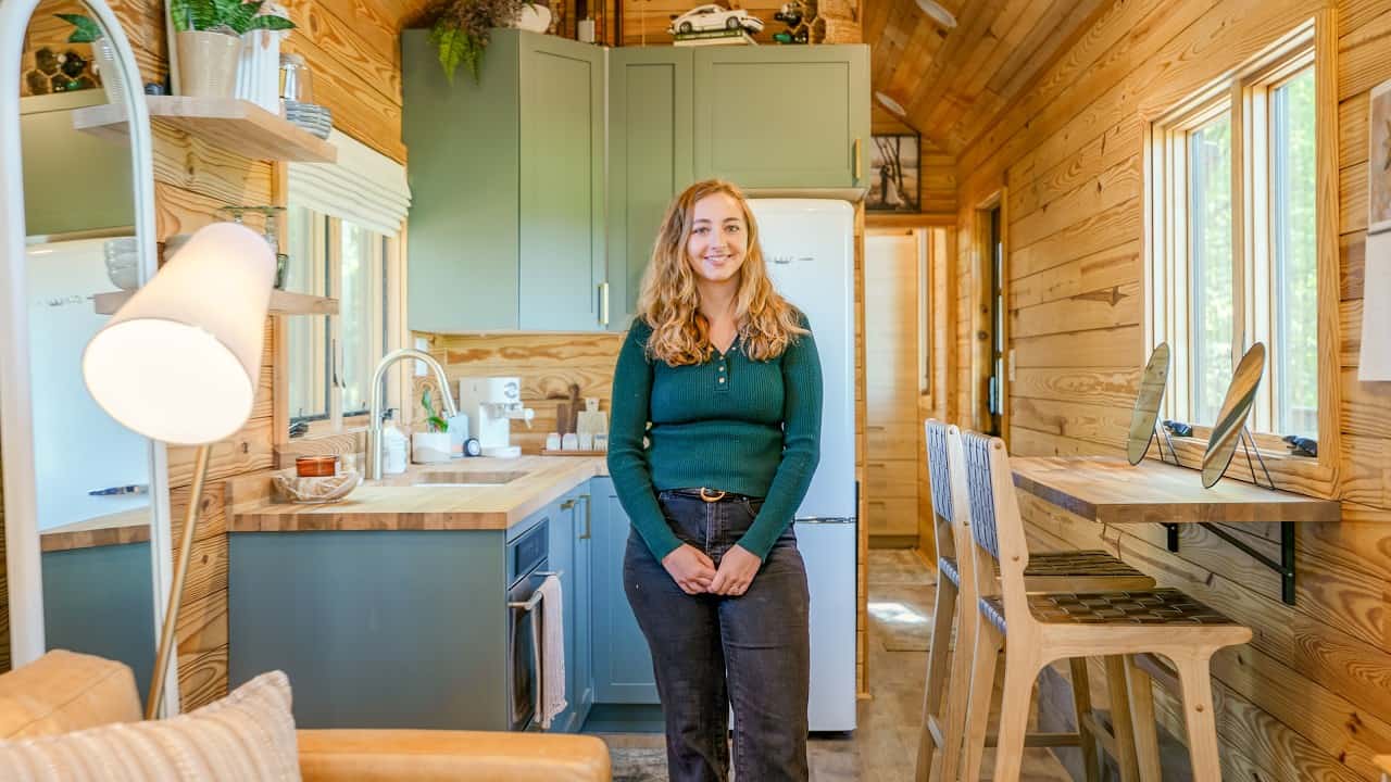 Single-story tiny cottage kitchen and living room, vaulted wood ceiling, sage cabinets, Mollie standing in the center