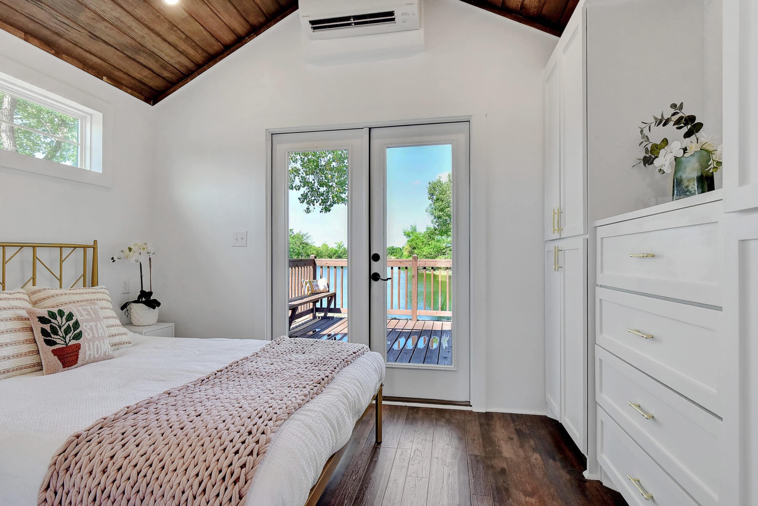 Bedroom with a gold-frame bed, white linens, a knit throw, and glass French doors opening onto a wooden deck over the lake at Monarch Springs Ranch