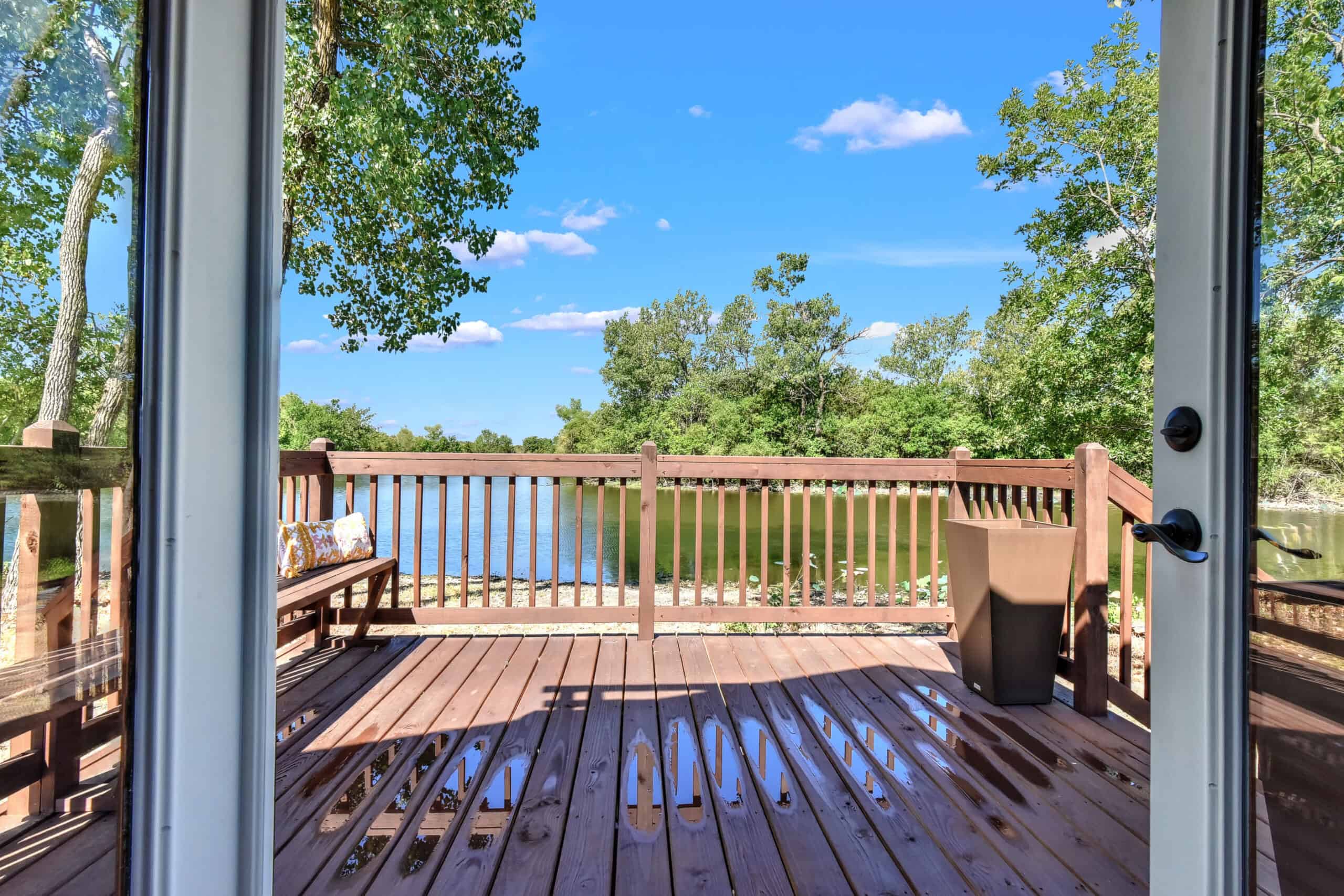 Open glass door onto a wet wooden deck with a bench cushion and a planter, looking over the private lake at Monarch Springs Ranch