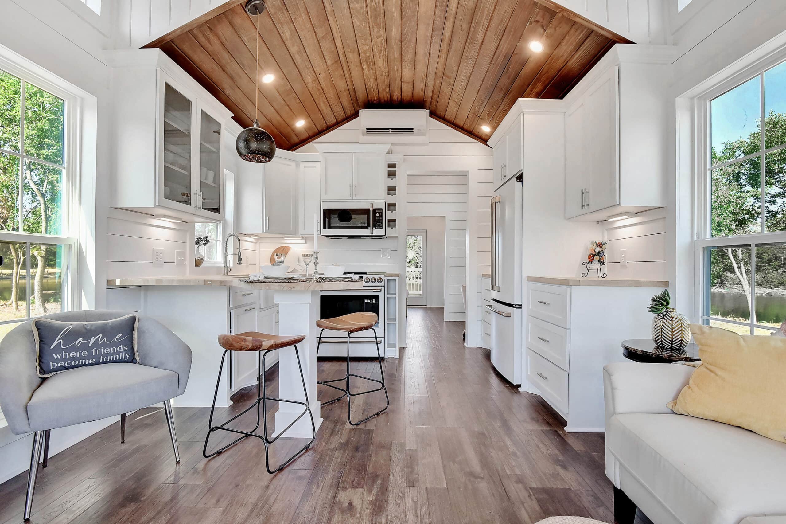 Open kitchen and living room with white shiplap, a wood-plank vaulted ceiling, a peninsula with two stools, and lake windows at Monarch Springs Ranch