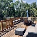 Wooden lakeside deck with a dining table and folded umbrella, a wicker lounge sofa, a bench along the rail, and the private lake through the trees at Monarch Springs Ranch in Celeste, Texas