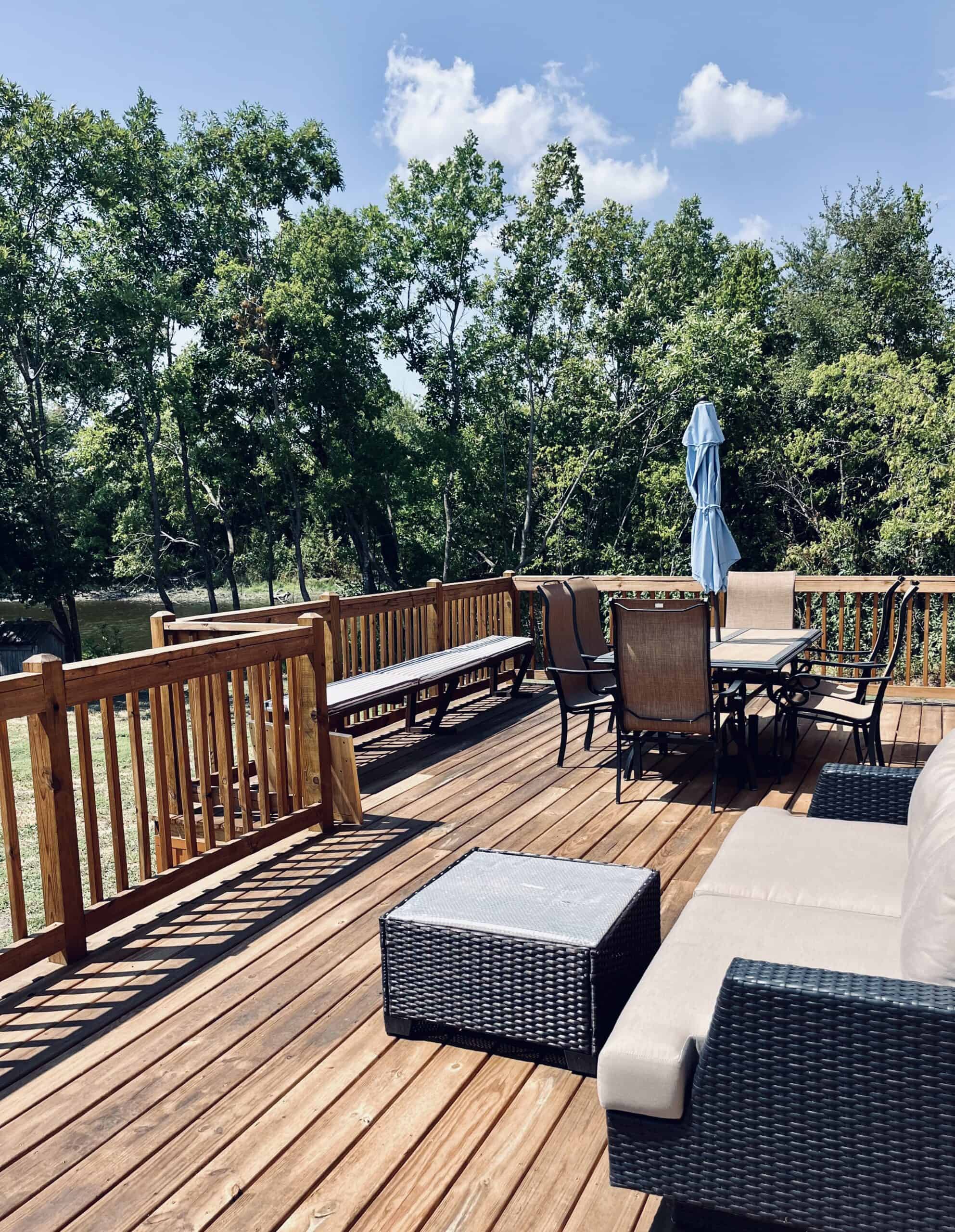Wooden lakeside deck with a dining table and folded umbrella, a wicker lounge sofa, a bench along the rail, and the private lake through the trees at Monarch Springs Ranch in Celeste, Texas