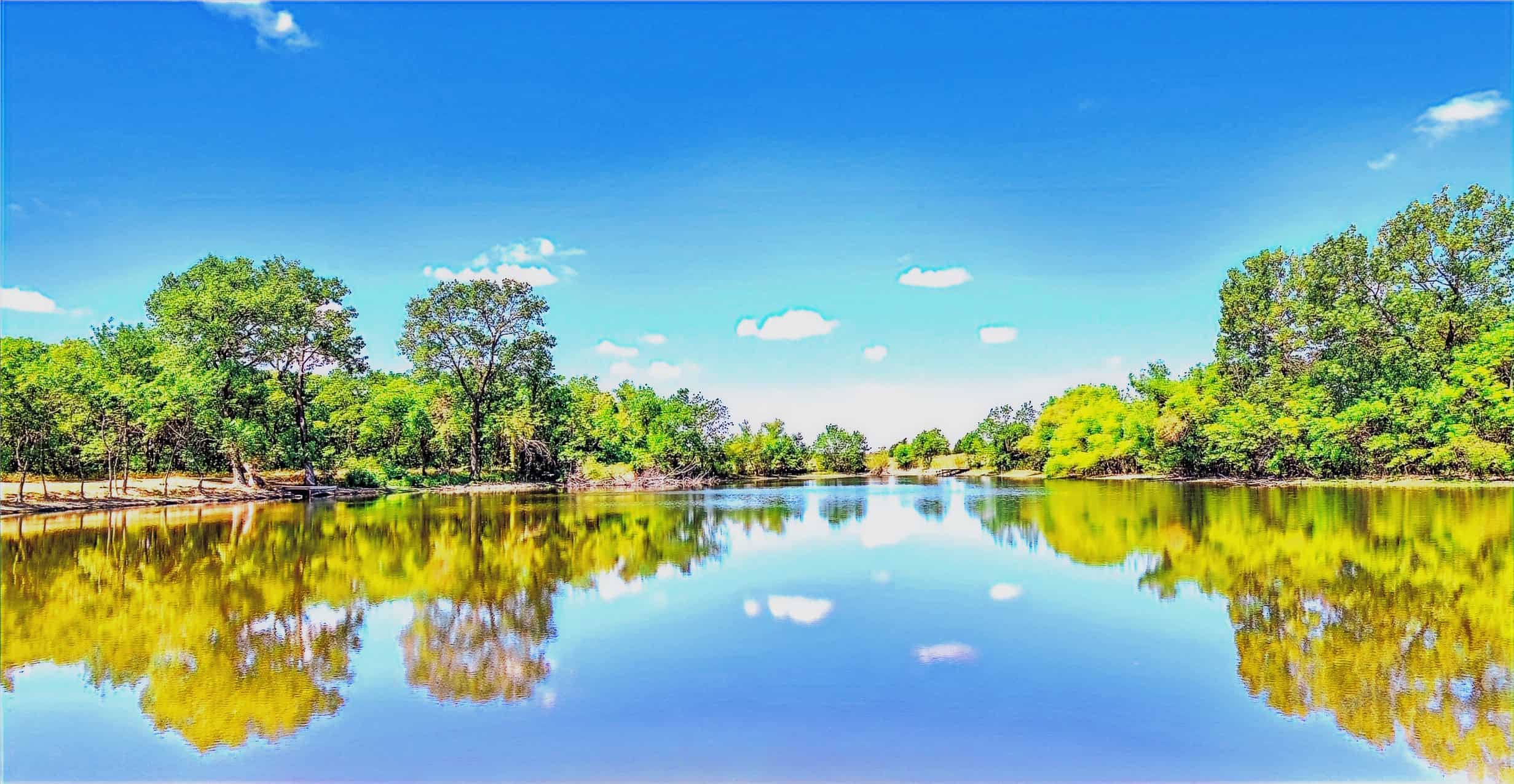 Still private lake ringed by green trees under a blue sky, with a small wooden dock on the left bank at Monarch Springs Ranch in Celeste, Texas