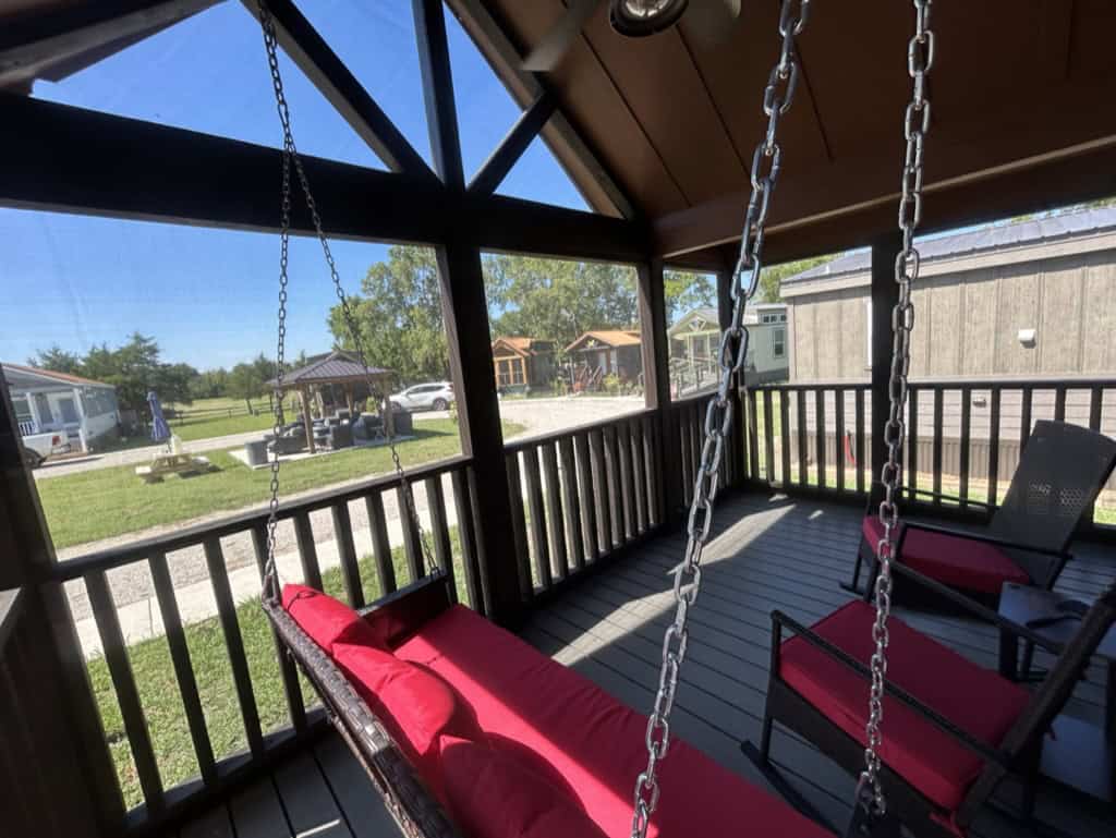 Screened porch with a red-cushion swing and rockers looking out at the green, a pergola, and neighboring cottages at Monarch Springs Ranch